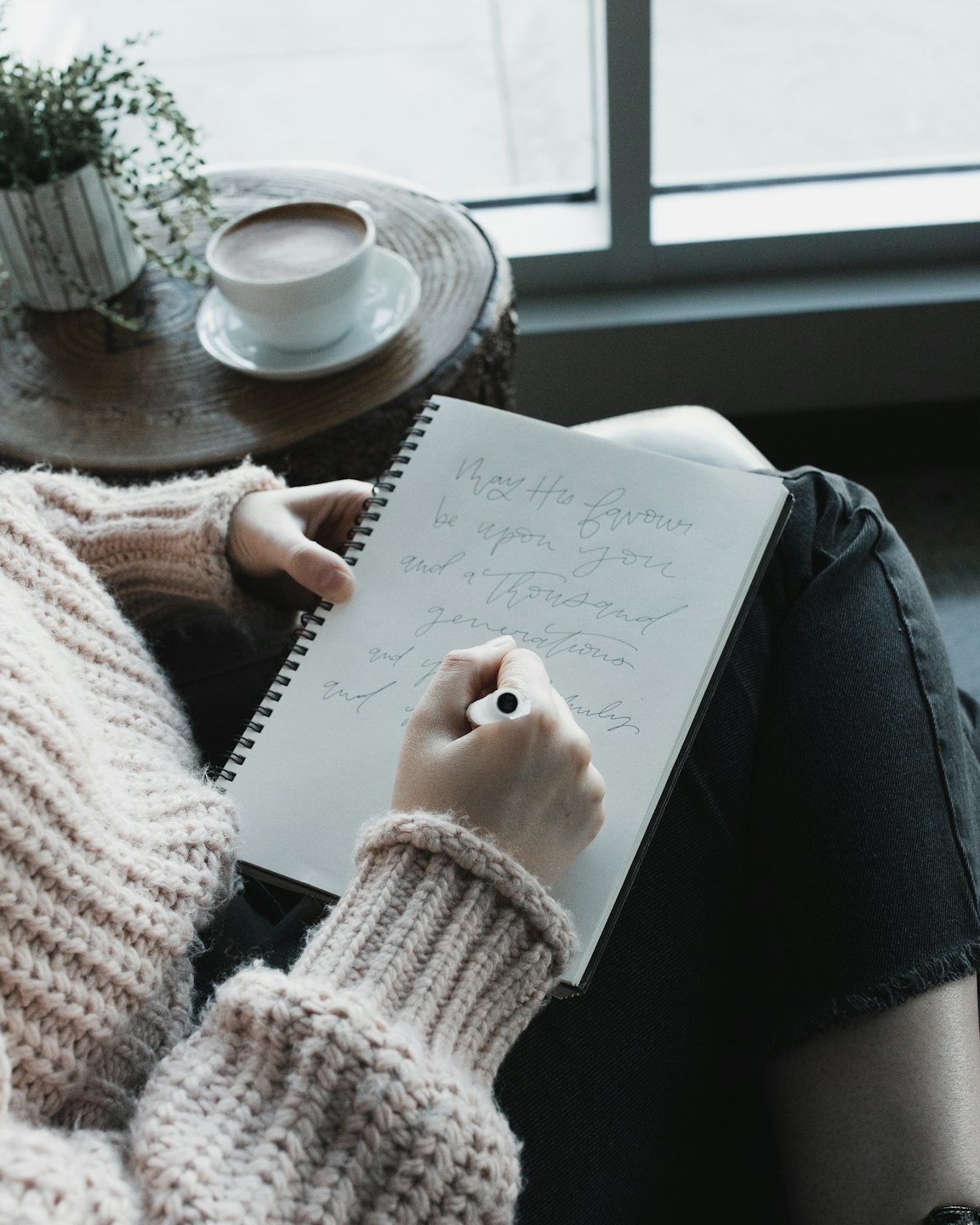 Person in beige sweater writing in notebook with pen.